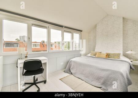 Helles und luftiges Schlafzimmer mit moderner Einrichtung, komfortablem Bett mit grauer Bettwäsche, weißem Schreibtisch mit schwarzem Stuhl und großen Fenstern Stockfoto