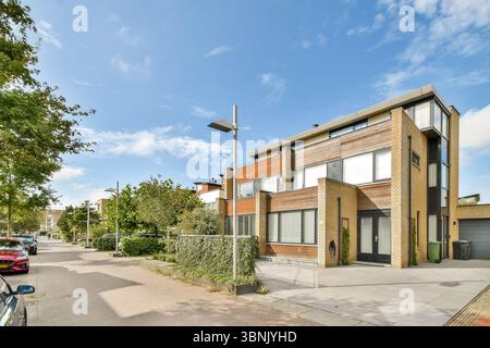 Ein modernes Vorstadthaus mit Ziegel- und Holzakzenten steht unter einem hellblauen Himmel in einer ruhigen Wohnstraße. Üppiges Grün und geparkte Autos säumen die Linie Stockfoto