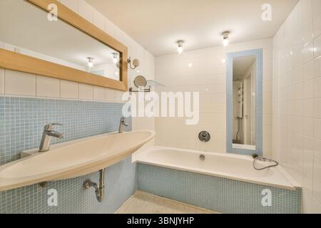 Ein modernes Badezimmer mit einem ovalen Waschbecken mit zwei Armaturen, einer geräumigen Badewanne und einem großen gerahmten Spiegel. Sanfte Beleuchtung ergänzt das saubere weiße A Stockfoto