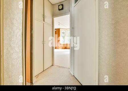Blick auf einen Flur mit hellen Wänden, Holzakzenten und einer Tür, die zu einem hellen Raum mit einem großen Fenster führt, das natürliches Licht im Inneren ermöglicht. Stockfoto