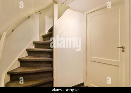 Eine gemütliche Inneneinrichtung mit einer geschwungenen Treppe mit Teppichboden neben einer geschlossenen weißen Tür. Das Ambiente strahlt eine schlichte und klare Ästhetik aus. Stockfoto