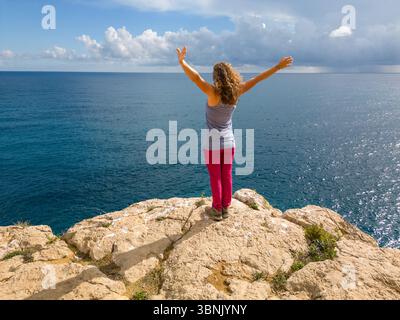 Eine junge Frau mit ausgestreckten Armen, die auf einer felsigen Klippe an der Costa Blanca, Spanien, steht. Das riesige blaue Meer erstreckt sich vor ihr unter einer teilweise Wolke Stockfoto