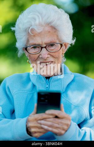 Eine ältere Frau mit weißem Haar und Brille lächelt, während sie ein Smartphone im Freien benutzt. Sie trägt eine hellblaue Fleecejacke und Appa Stockfoto