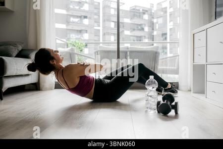 Eine Frau trainiert zu Hause und macht Übungen auf einem Holzboden. Sie benutzt Kurzhanteln und hat eine Flasche Wasser in der Nähe, mit hellem Tageslicht flo Stockfoto