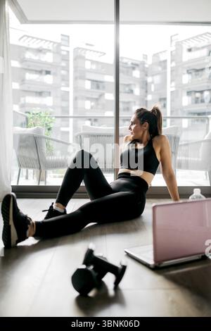 Eine Frau in schwarzer Sportswear macht eine Pause von ihrem Heimtraining und sitzt auf dem Boden ihrer Wohnung. In der Nähe deuten ein Laptop und Kurzhanteln auf ihre exe an Stockfoto
