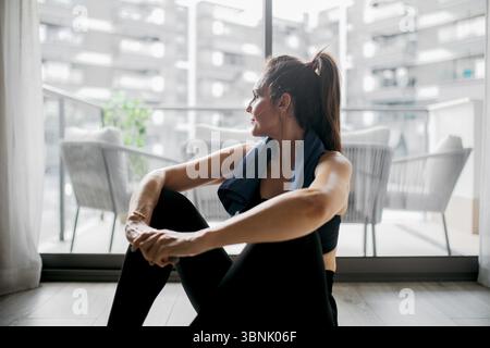 Eine Frau nimmt sich einen Moment Zeit, um sich nach ihrem Workout zu Hause zu entspannen und sitzt mit einem Handtuch um den Hals auf dem Boden. Sie blickt nach draußen und denkt über ihn nach Stockfoto
