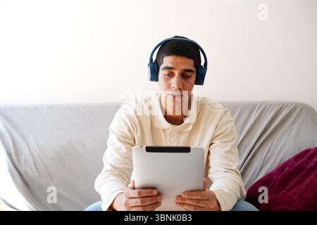 Ein junger arabischer Mann sitzt auf einer Couch, trägt Kopfhörer und benutzt ein Tablet. In seine Routine eingetaucht, ist er konzentriert und entspannt und genießt einen Moment digi Stockfoto