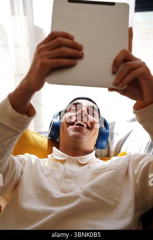 Ein junger arabischer Mann entspannt sich auf einer bequemen Couch, lächelt und engagiert sich mit seinem Tablet. Mit blauen Kopfhörern genießt er seine tägliche Routine der digitalen Medien Stockfoto