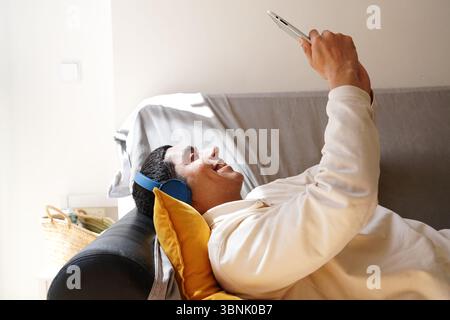 Ein junger arabischer Mann, der auf einem Sofa liegt und blaue Kopfhörer trägt, benutzt ein Tablet in entspannter Atmosphäre. Er lächelt, während er seine Freizeit genießt und Stockfoto