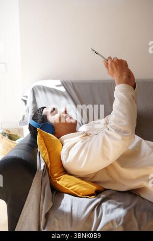 Ein junger arabischer Mann, der auf einem Sofa liegt und blaue Kopfhörer trägt, benutzt ein Tablet in entspannter Atmosphäre. Er lächelt, während er seine Freizeit genießt und Stockfoto