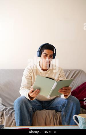 Ein junger arabischer Mann sitzt bequem auf einem Sofa, trägt Kopfhörer und liest ein Buch. Er scheint entspannt zu sein und genießt einen Moment der Freizeit in seiner täglichen Rutte Stockfoto