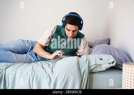 Ein junger arabischer Mann entspannt sich auf seinem Bett, während er blaue Kopfhörer trägt und ein Tablet benutzt. Er scheint konzentriert zu sein und genießt seine tägliche Routine in einer lässigen und c Stockfoto
