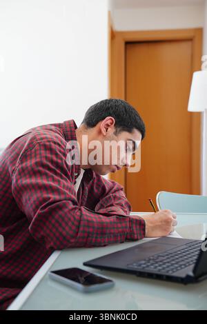 Ein junger arabischer Mann konzentriert sich auf das Schreiben an einem Tisch, umgeben von einem Laptop und einem Smartphone. Die Szene fängt einen Moment aus seiner täglichen Routine ein, Stockfoto