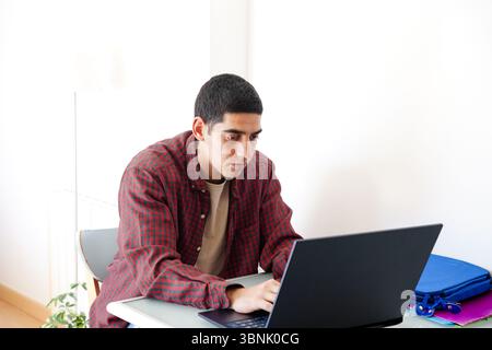 Junger arabischer Mann, der an einem Tisch in einem hellen Raum sitzt und sich darauf konzentriert, an seinem Laptop zu arbeiten. Die Szene zeigt einen typischen Tag in seinem Leben und hebt die Moderne hervor Stockfoto