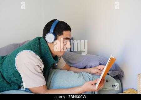 Ein junger arabischer Mann liegt bequem auf seinem Bett, trägt Kopfhörer und genießt es, ein Buch zu lesen. Die Szene fängt einen friedlichen Moment in seinem täglichen Ro ein Stockfoto