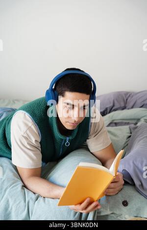 Ein junger arabischer Mann entspannt sich auf seinem Bett und liest in der Freizeit, während er blaue Kopfhörer trägt. Er ist bequem gekleidet und in seinen Alltag eingetaucht Stockfoto