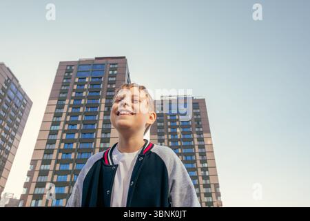 Ein fröhlicher Junge mit einem großen Lächeln steht vor hohen Wolkenkratzern und fängt an einem hellen, sonnigen Tag das Wesen des Stadtlebens ein. Die Szene strahlt Sie aus Stockfoto