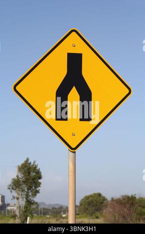 Gelbes, geteiltes Straßenschild an einem Metallpfosten vor einem klaren blauen Himmel Stockfoto