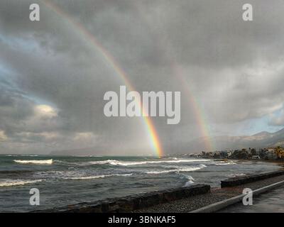 Ein spektakulärer Doppelregenbogen über dem stürmischen Mittelmeer an der Nordküste Kretas, Griechenland. Wellen krachen am Ufer unter dramatischem Himmel. Stockfoto