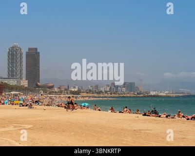 Am beliebten Strand Barceloneta mit der modernen Skyline der Stadt, darunter das Hotel Arts und der Mapfre Tower, entspannen sich die Menschen am Strand. Stockfoto