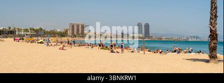 Ein Panoramablick auf einen sonnenverwöhnten Strand von Barcelona, voller Sonnenanbeter und Schwimmer, mit den berühmten Doppeltürmen von Port Olímpic in der Ferne. Stockfoto