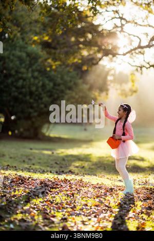 Südasiatische Mädchen mit Feenflügeln, die Sternstab und Orangenbox auf blattbedeckter Rodung halten Stockfoto
