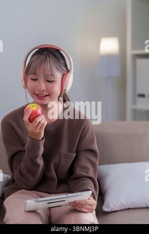 Strahlende junge asiatische Frau mit Kopfhörern entspannt sich auf der Couch, genießt einen frischen Apfel und stöbert auf einem Tablet, Freizeit in Innenräumen mit Technologie und F Stockfoto