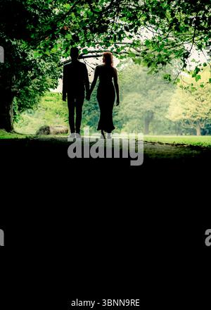 Ein Paar geht Hand in Hand auf einem Weg in einem Park, der sich vor dem Licht abhebt. Stockfoto