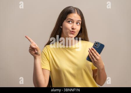 Eine nachdenkliche junge Frau, die ein Smartphone hält, schaut zur Seite und zeigt, als ob sie verschiedene Optionen im Internet in Betracht ziehen oder wählen würde. Stockfoto