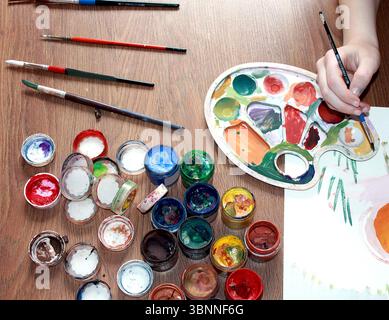 Ein Mädchen malt mit Farbe. Nahaufnahme einer Hand. Gouache, Pinsel und Papierblätter auf hölzernem Hintergrund. Stockfoto