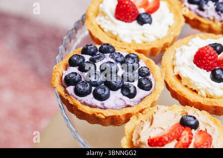Mini-Obstkuchen mit Sahne und Beeren. Buntes Dessert mit Heidelbeeren und Erdbeeren. Süßes Gebäck auf Glasplatte. Leckeres Vergnügen zum Feiern Stockfoto