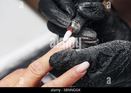 Nageltechniker feilt Fingernägel. Nahaufnahme der Hände in Handschuhen und Kundenhand. Maniküre mit dem elektrischen Nagelwerkzeug. Schönheitsbehandlung im Salon Stockfoto