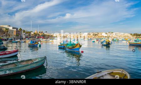 Marsaxlokk, Malta - 9. Februar 2024: Panoramablick auf das farbenfrohe Luzzu, traditionelle Fischerboote bei Wintersonnenuntergang mit dem Dorf Marsaxlokk im bac Stockfoto