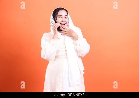 Eine junge muslimische Frau in einem Hijab spricht mit einem Ausdruck der Überraschung und Freude an ihrem Telefon und zeigt eine positive Emotion, die auf einer Orange isoliert ist Stockfoto