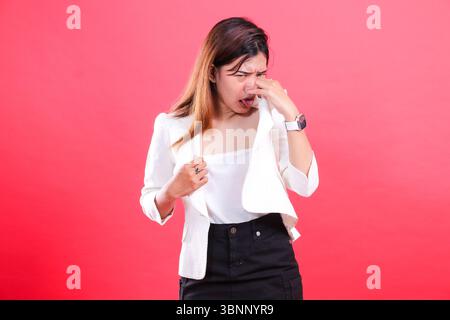 Eine Frau, die eine weiße Jacke und eine schwarze Hose trägt, hält ihre Nase und streckt ihre Zunge heraus, isoliert auf einem roten Hintergrund Stockfoto