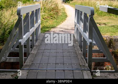 Hölzerne Fußgängerbrücke über einen kleinen Kanal mit einem Kiesweg umgeben von üppigem grünem Gras und Sommervegetation Stockfoto