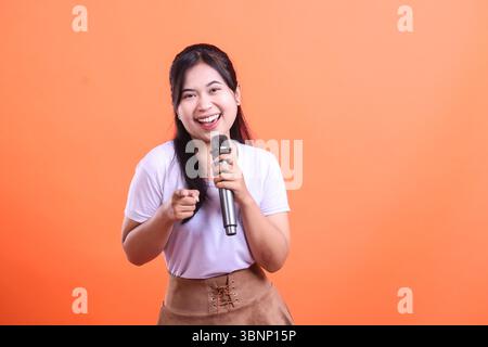 Eine junge asiatische Frau lächelt glücklich, während sie ein Mikrofon hält und mit einer positiven Einstellung auf ihren Finger zeigt, isoliert auf orangefarbenem Hintergrund Stockfoto