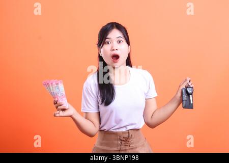 Eine glückliche asiatische Frau, die Geld und Autoschlüssel hält, sieht überrascht und aufgeregt aus. Das bedeutet, dass sie das Auto mit Bargeld auf orangem Hintergrund gewonnen hat. Stockfoto