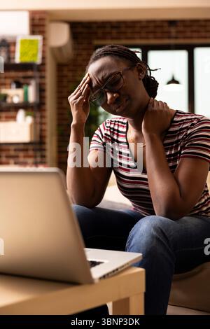 Schwarze Unternehmerin lehnt sich auf das Knie, frustriert bei der Arbeit an der Marketingpräsentation auf dem Notebook. afroamerikanische Frau mit geschlossenen Augen, die sich von der Arbeit überwältigt fühlt und Kopfschmerzen hat. Stockfoto