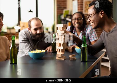 Junger schwarzer Mann mit Brille, der während der Spielsitzung in der Halle gerne Holzblock vom Jenga-Turm zieht. Verschiedene Gruppen von Freunden beobachten aufmerksam und genießen gemeinsame Momente in einem gemauerten Raum. Stockfoto