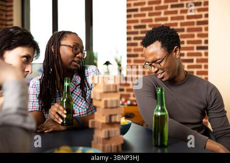 afroamerikanisches Paar trinkt Bier und macht im Mauerraum dumme Gesichtsausdrücke. Gruppe von Freunden am Tisch, genießen Brettspiele und entspannte Bonding-Zeit mit Holzspielstücken. Stockfoto