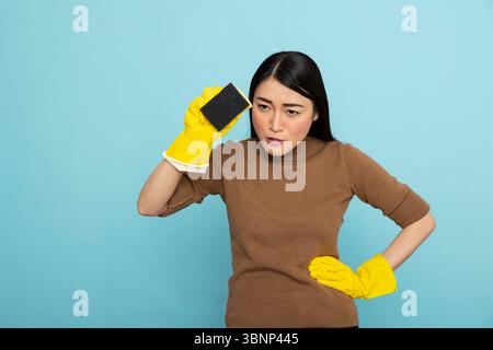 Porträt einer seriösen asiatischen Frau in gelben Gummihandschuhen, die Reinigungsschwamm und die Hand auf der Taille hält und mit Absicht nach vorne starrt. Hausmädchen, das selbstbewusst und präzise auf die Arbeit vorbereitet ist. Stockfoto
