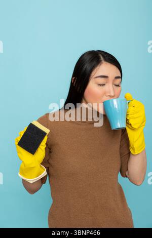 Asiatische Hausangestellte schlürfen friedlich Kaffee, während sie einen Schwamm halten und während der Hausarbeiten eine entspannende Pause einlegen. Weibliche Hausmeisterin mit gelben Gummihandschuhen, genießt während der Frühjahrsputz einen ruhigen Moment. Stockfoto