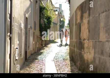 Zwei Personen laufen Hand in Hand durch eine malerische Kopfsteinpflasterallee in Cascais, Portugal. Das warme Sonnenlicht strahlt ein sanftes Licht aus und unterstreicht die malerische Umgebung. Stockfoto