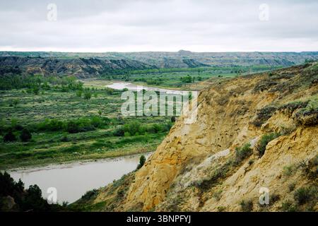 Der Little Missouri River schlängelt sich durch grüne Ebenen und geschnitzte Klippen im Theodore Roosevelt National Park. Bewölkter Himmel macht die Szene weicher. Stockfoto
