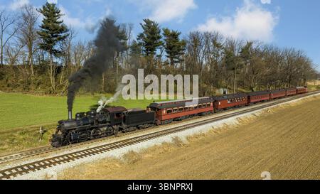Ein Blick auf die Drohne eines restaurierten Dampflokomotivzugs, der durch Farmland reist und an einem Herbsttag zu einem kleinen Bahnhof fährt Stockfoto