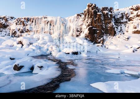 Winterszene im Freien mit einem schneebedeckten Fluss in der Nähe des teilweise gefrorenen Oxararfoss Wasserfalls im Thingvellir Nationalpark im Goldenen Kreis in Island. Stockfoto