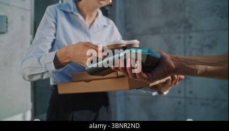 Liefermann übergibt Geschäftsfrau Zahlungsterminal nach erfolgreicher Pizzalieferung zum Bürogebäude. Frau hält Essen aus dem Restaurant, bezahlt für Bestellung kontaktlos mit Smartphone. Nahaufnahme. Stockfoto