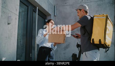 Liefermann auf Elektroroller brining Papiertüte mit Online-Einkaufsauftrag für Geschäftsfrau, die im Büro arbeitet. Frau, die während des Arbeitstages das Paket vom Food Market Delivery Service nimmt. Zeitlupe Stockfoto