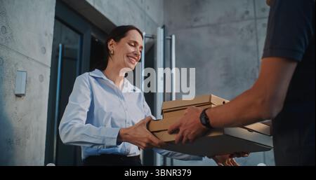 Mann, der im Pizzalieferdienst arbeitet, Restaurant-Essen für Kunden, die in einem modernen Bürogebäude arbeiten. Schöne Geschäftsfrau, die Pizza in Kartons vom Liefermann nimmt. Zeitlupe. Stockfoto
